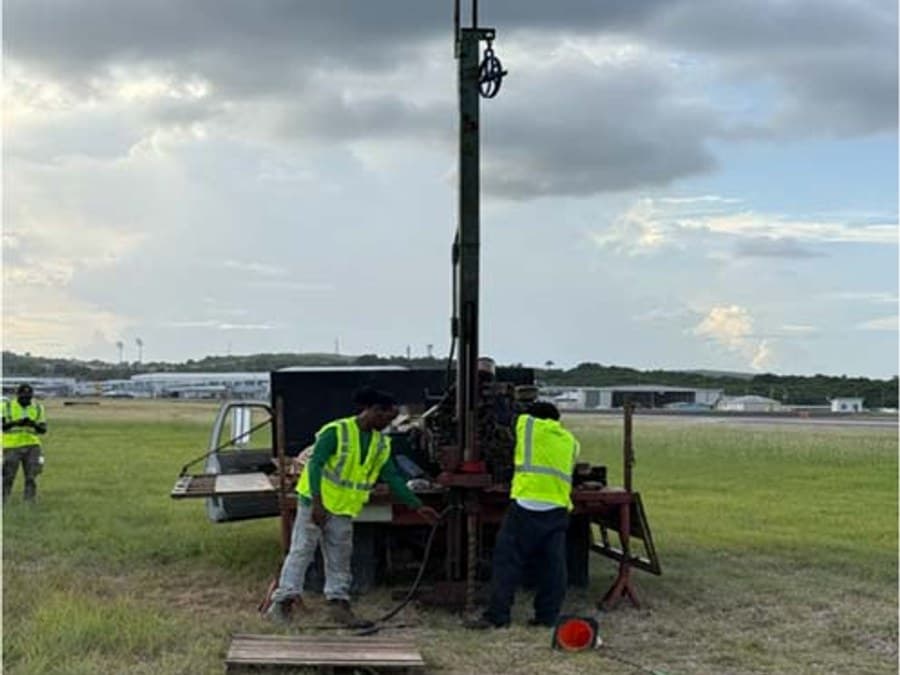 GID field crew operating a rock coring rig on the airfield at V.C. Bird International Airport, with the runway visible behind